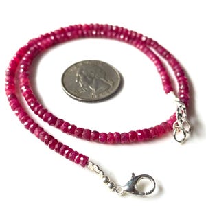 May include: A vibrant ruby gemstone necklace with a silver clasp and a quarter coin for size reference. The necklace is composed of small, faceted, deep red gemstones strung together, creating a delicate and elegant accessory.