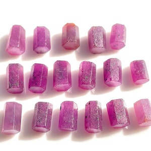 Può includere: Una collezione di 18 gemme di rubino rosa sfaccettate, ciascuna con una forma a barilotto esagonale. Le gemme sono disposte in tre file di sei, con ogni fila leggermente sfalsata rispetto a quella sopra. Le gemme sono tutte di dimensioni e forma simili e hanno una finitura liscia e lucida.