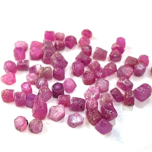 May include: A collection of pink ruby gemstones, each with a faceted, rough-cut shape. The rubies are scattered on a white surface.