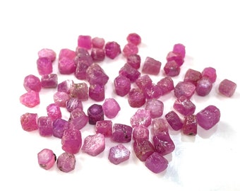 Può includere: Una collezione di pietre preziose di rubino rosa, ciascuna con una forma sfaccettata e grezza. I rubini sono sparsi su una superficie bianca.