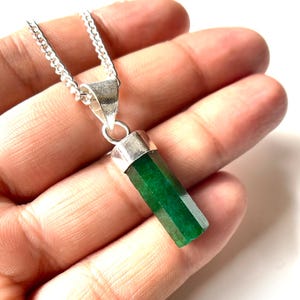 Puede incluir: Un collar de plata con un colgante de piedra preciosa verde esmeralda facetada. El colgante es rectangular con lados angulados. La cadena es una delicada cadena de plata. La piedra preciosa es de un color verde intenso y vibrante.