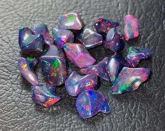 Può includere: Una collezione di pietre preziose di opale nero lucido. Gli opali mostrano vibranti lampi di colore, tra cui rosso, verde e blu, contro una superficie scura e lucida. Le pietre variano per dimensioni e forma, con una finitura liscia e riflettente.