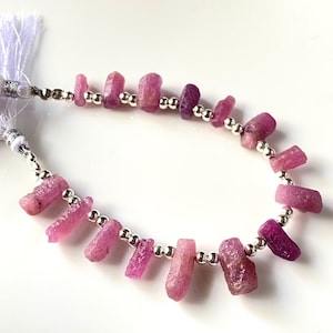 Op de afbeelding: Een streng roze en paarse ruwe edelsteen kralen geregen op een zilveren ketting met een witte kwast aan elk uiteinde.