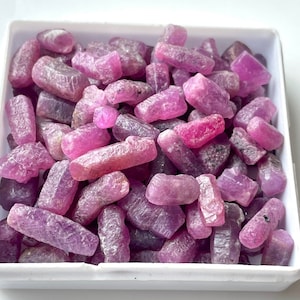 May include: A collection of pink ruby rough stones in a white box. The stones are various sizes and shapes, with a rough, natural finish.