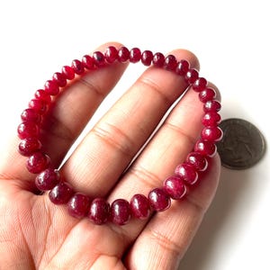 Puede incluir: Un brazalete de cuentas compuesto por pequeñas piedras preciosas redondas de color rojo intenso. El brazalete se sostiene en una mano, con una moneda de plata visible en el fondo. Las piedras tienen un aspecto pulido y brillante. El brazalete mide unos 18 cm de circunferencia.