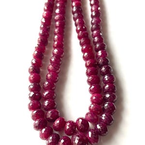 Può includere: Tre fili di perline di pietre preziose rosse scuro sfaccettate. Le perline sono infilate insieme, creando un aspetto simile a una collana. Le pietre preziose hanno una superficie lucida e riflettente, su sfondo bianco.