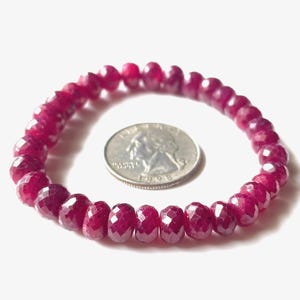 May include: A ruby gemstone bracelet with faceted beads in shades of deep red and pink. A silver coin is next to the bracelet for size comparison. The bracelet is on a white background.