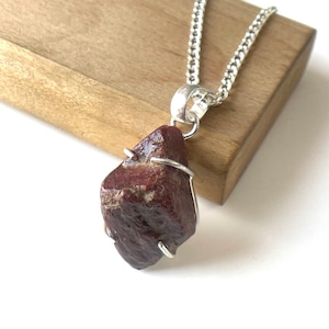 May include: A raw, dark red gemstone pendant with a silver-colored metal wire frame and chain. The irregularly shaped stone is suspended from a silver-toned bail. The necklace rests on a wooden block, showcasing the natural texture and color of the stone.