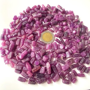 Puede incluir: Un montón grande de pequeños cristales de rubí púrpura sin pulir. Los cristales son de varias formas y tamaños, algunos son largos y delgados, mientras que otros son más redondeados. Se incluye una moneda pequeña para la escala.