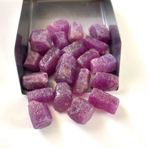 Puede incluir: Una colección de piedras preciosas de rubí en bruto, sin pulir, de color rojo rosáceo, con formas cristalinas irregulares y superficies texturizadas, contenidas dentro de una pala de metal de tono plateado.