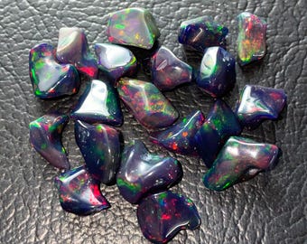 May include: A collection of polished black opal gemstones. Each stone displays vibrant flashes of color, including red, green, and blue, against a dark, glossy surface. The opals vary in size and shape, with a smooth, rounded appearance.