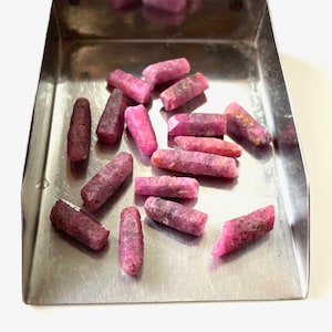 May include: A collection of pink ruby crystals in a metal tray. The crystals are all pointed and have a rough, natural texture.