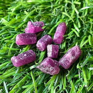 Puede incluir: Una colección de siete pequeñas piedras preciosas de color púrpura ásperas sobre un fondo de hierba artificial verde.