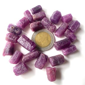 May include: A collection of small, rough, purple ruby crystals. The crystals are arranged in a circle around a Thai baht coin.