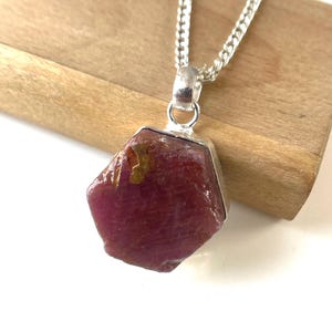 May include: A ruby pendant necklace with a silver chain. The ruby is a deep red color with a rough, natural texture. The pendant is set in a silver bezel. The necklace is displayed on a wooden surface.