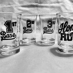 Puede incluir: Cuatro vasos de chupito de vidrio transparente con letras negras. Los vasos están etiquetados con "1st Base", "2nd Base", "3rd Base" y "Home Run".