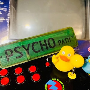 May include: A green wooden sign with the text "PSYCHO PATH" in black lettering. The sign is sitting on a black surface with red buttons and a yellow rubber duck with a blue beak.