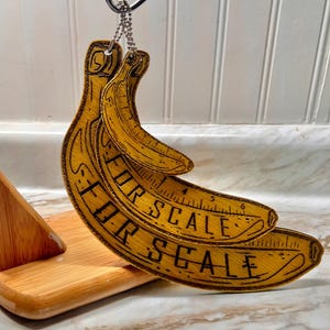 May include: Wooden banana-shaped measuring tools with the text "FOR SCALE" etched on them. The bananas are yellow with dark brown etched details, and are hanging from a metal hook. The measuring tools have inch markings.
