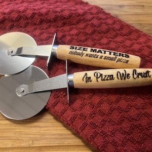 May include: Two pizza cutters with wooden handles. The top handle has the text "SIZE MATTERS nobody wants a small pizza" engraved on it. The bottom handle has the text "In Pizza We Crust" engraved on it.