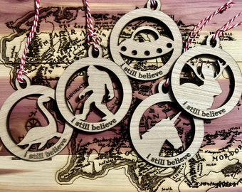 May include: Five wooden ornaments with a rustic brown finish. Each ornament features a different silhouette: a UFO, a Bigfoot, a deer, a unicorn, and a goose. Each ornament has the text "I still believe" inscribed on it.