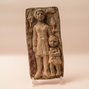 May include: A rectangular, beige-colored relief sculpture depicting a woman and a child. The figures are carved in a simple, stylized manner, with the woman standing and the child in front of her. The sculpture is mounted on a clear stand.