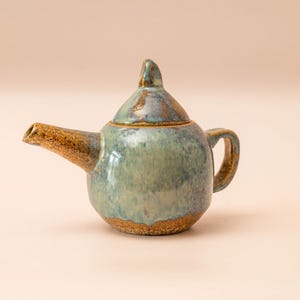 Può includere: Piccola teiera in ceramica fatta a mano, con corpo arrotondato e coperchio. Smaltata in tonalità blu e verde, con beccuccio, manico e base marroni. Design semplice ed elegante, adatta per servire il tè.