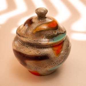Può includere: Una zuccheriera in ceramica con coperchio. La ciotola è rotonda con una superficie strutturata ed è decorata con colori marrone, arancione e verde acqua. Il coperchio ha un piccolo pomello sopra.
