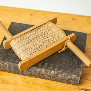 Op de afbeelding: Een houten spoel met een lengte van touw eromheen gewikkeld. De spoel ligt op een vintage boek met een donkerbruine kaft en gouden letters.