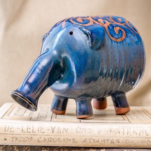 Può includere: Un salvadanaio di ceramica blu a forma di elefante con una lunga proboscide e disegni a spirale marroni. Il salvadanaio è seduto sopra due libri.