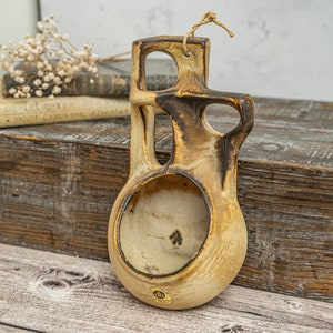 May include: A brown and beige ceramic wall planter with a unique abstract design. The planter has a hole in the back for hanging and a small opening in the front for planting.