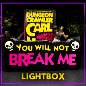 May include: A black lightbox with the text "YOU WILL NOT BREAK ME" in pink and "LIGHTBOX" in yellow. The lightbox has skull designs on each end. A book titled "Dungeon Crawler Carl" is behind the lightbox.