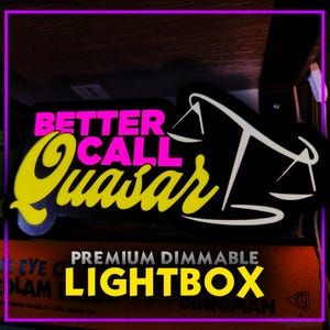May include: A black lightbox with the text "BETTER CALL QUASAR" in pink and yellow. The design includes a white scale icon. The words "PREMIUM DIMMABLE LIGHTBOX" are in yellow below the main text.