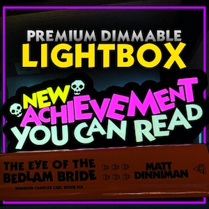 May include: A black lightbox with the text "PREMIUM DIMMABLE LIGHTBOX" in yellow and white. Below, a neon sign reads "NEW ACHIEVEMENT YOU CAN READ" with skull graphics. The book "THE EYE OF THE BEDLAM BRIDE" is visible below.