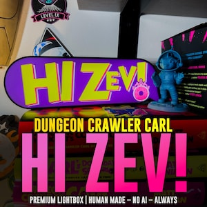 May include: A rectangular purple lightbox featuring the bright yellow text HI ZEV! with a pink donut icon. Next to it stands a small blue figurine of a character in a space suit. The lightbox rests on a stack of books titled Dungeon Crawler Carl.