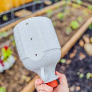 May include: A light gray, textured garden watering can with three holes on top and a small handle. The base is orange and black. The watering can is held in a hand, with a garden bed in the background.