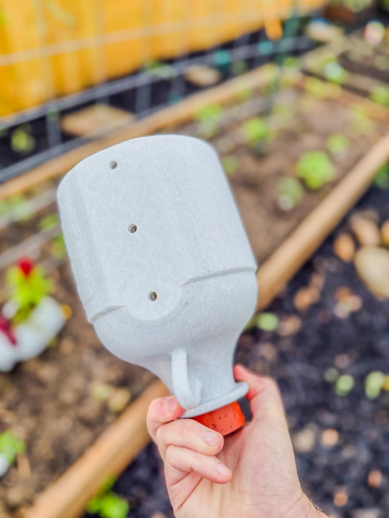 May include: A light gray, textured garden watering can with three holes on top and a small handle. The base is orange and black. The watering can is held in a hand, with a garden bed in the background.