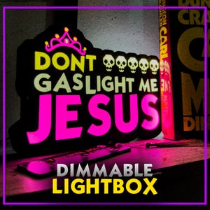 May include: A black dimmable lightbox with the text "DON'T GASLIGHT ME JESUS" in yellow and pink. The design includes a pink crown and skulls. The words "DIMMABLE LIGHTBOX" are at the bottom. A book is visible in the background.