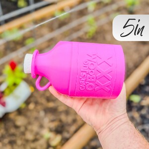 May include: A bright pink jug with a white cap and a handle. The jug has the text "Carl's Jug O'Book" and "XXX" printed on it. The jug is approximately 5 inches in height.