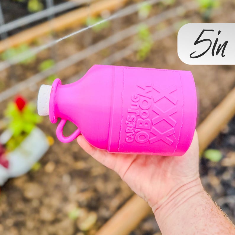 May include: A bright pink jug with a white cap and a handle. The jug has the text "Carl's Jug O'Book" and "XXX" printed on it. The jug is approximately 5 inches in height.