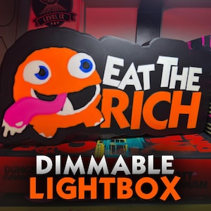 May include: A black rectangular lightbox featuring an orange cartoon character with large white eyes, blue pupils, and a long pink tongue. The text EAT THE RICH is displayed in bold white and orange lettering against the black background.