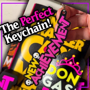 May include: A black keychain with the word "ACHIEVEMENT" in pink, and "NEW ACHIEVEMENT" with skull graphics. The keychain is held in a hand, with the text "The Perfect Keychain!" in the background.
