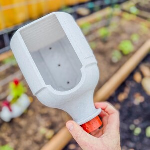 May include: A light gray, rectangular planter with a textured surface and an orange base. The planter is held over a garden bed with visible plants and a wooden handle. The planter is designed for gardening.