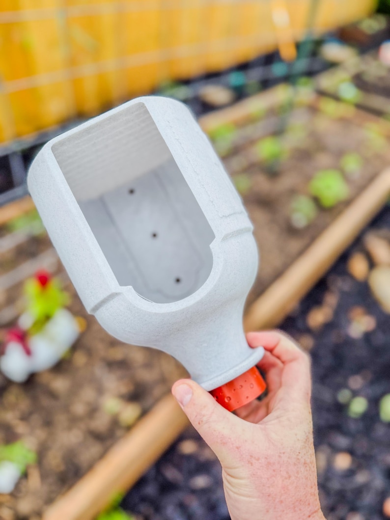 May include: A light gray, rectangular planter with a textured surface and an orange base. The planter is held over a garden bed with visible plants and a wooden handle. The planter is designed for gardening.