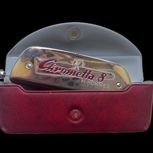 May include: A Chrometta 8 M. Hohner harmonica in a red case with a gray lid. The harmonica is silver and gold with the name "Chrometta 8" in red script. The case is rectangular with a snap closure.