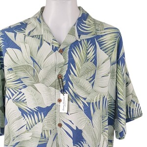Puede incluir: Una camisa de manga corta con botones azul y verde con estampado de hojas tropicales. La camisa tiene cuello y cierre de botones.