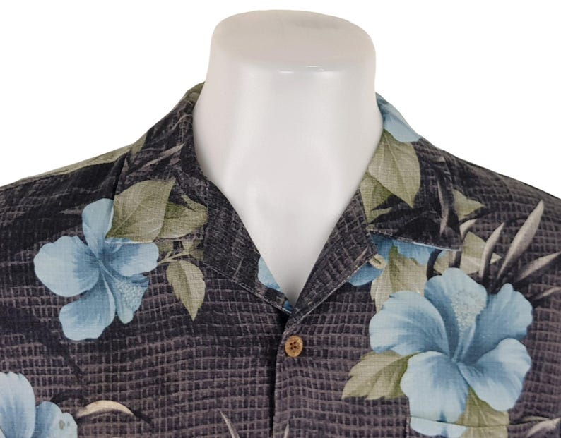 K&ouml;nnte beinhalten: Ein schwarzes Hemd mit Kn&ouml;pfen und einem blauen und gr&uuml;nen Blumenmuster. Das Hemd hat einen Kragen und einen Knopfverschluss.