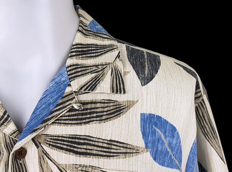May include: A beige button-up shirt with a tropical leaf print in shades of blue, black, and brown. The shirt has a collar and a button closure.