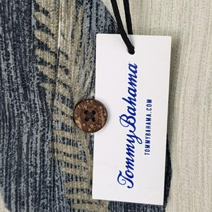 May include: A white tag with the text "Tommy Bahama" in blue lettering and the website address "tommybahama.com" printed below. The tag is attached to a light beige fabric with a blue and brown patterned design.