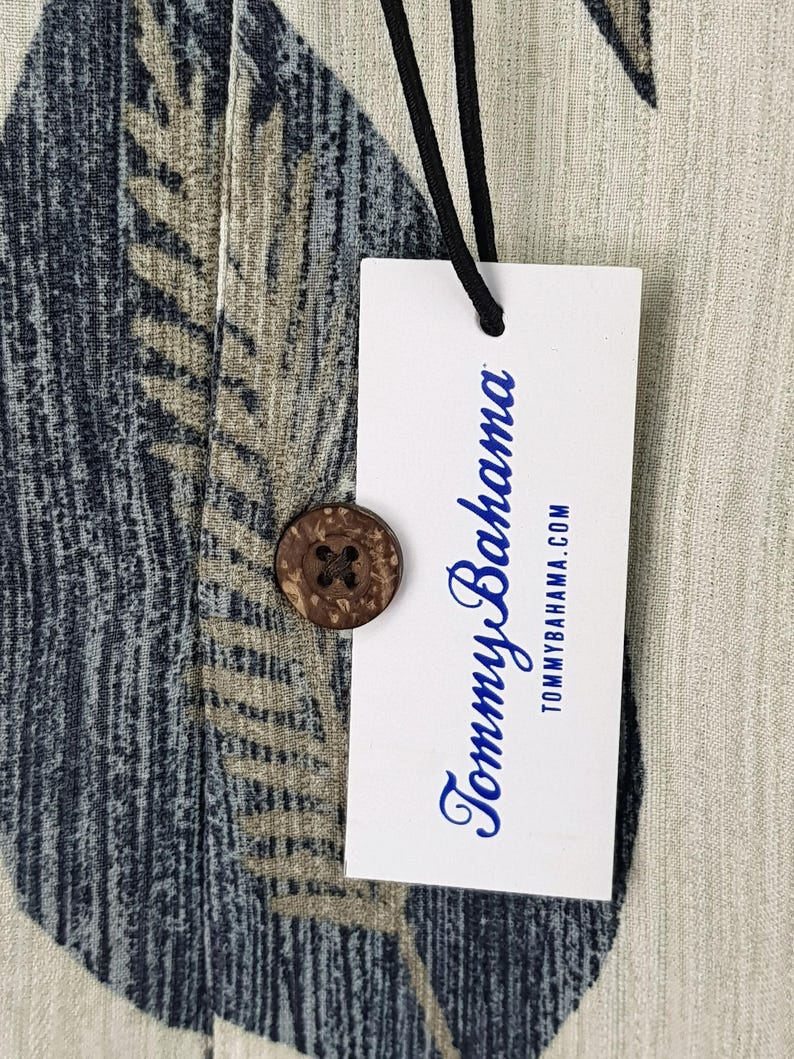 May include: A white tag with the text "Tommy Bahama" in blue lettering and the website address "tommybahama.com" printed below. The tag is attached to a light beige fabric with a blue and brown patterned design.