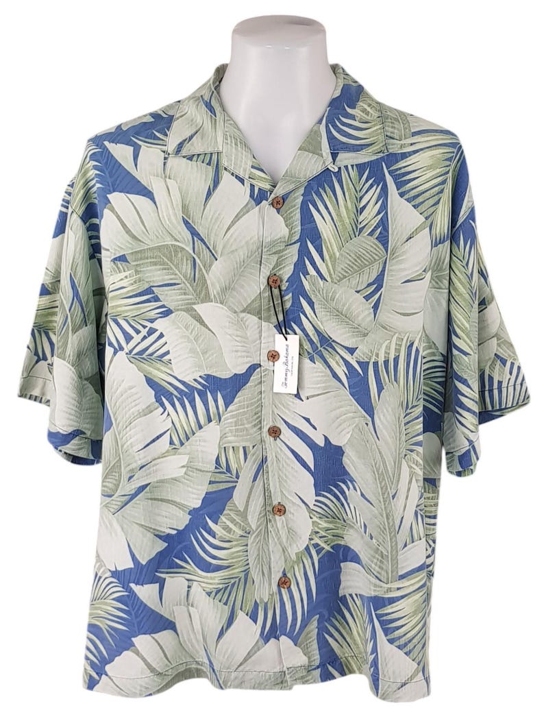 Puede incluir: Una camisa de manga corta con botones azul y verde con estampado de hojas tropicales. La camisa tiene cuello y cierre de botones.